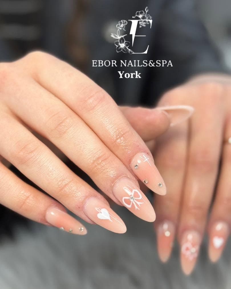 Nude almond nails with hand-painted white bows, hearts and gems